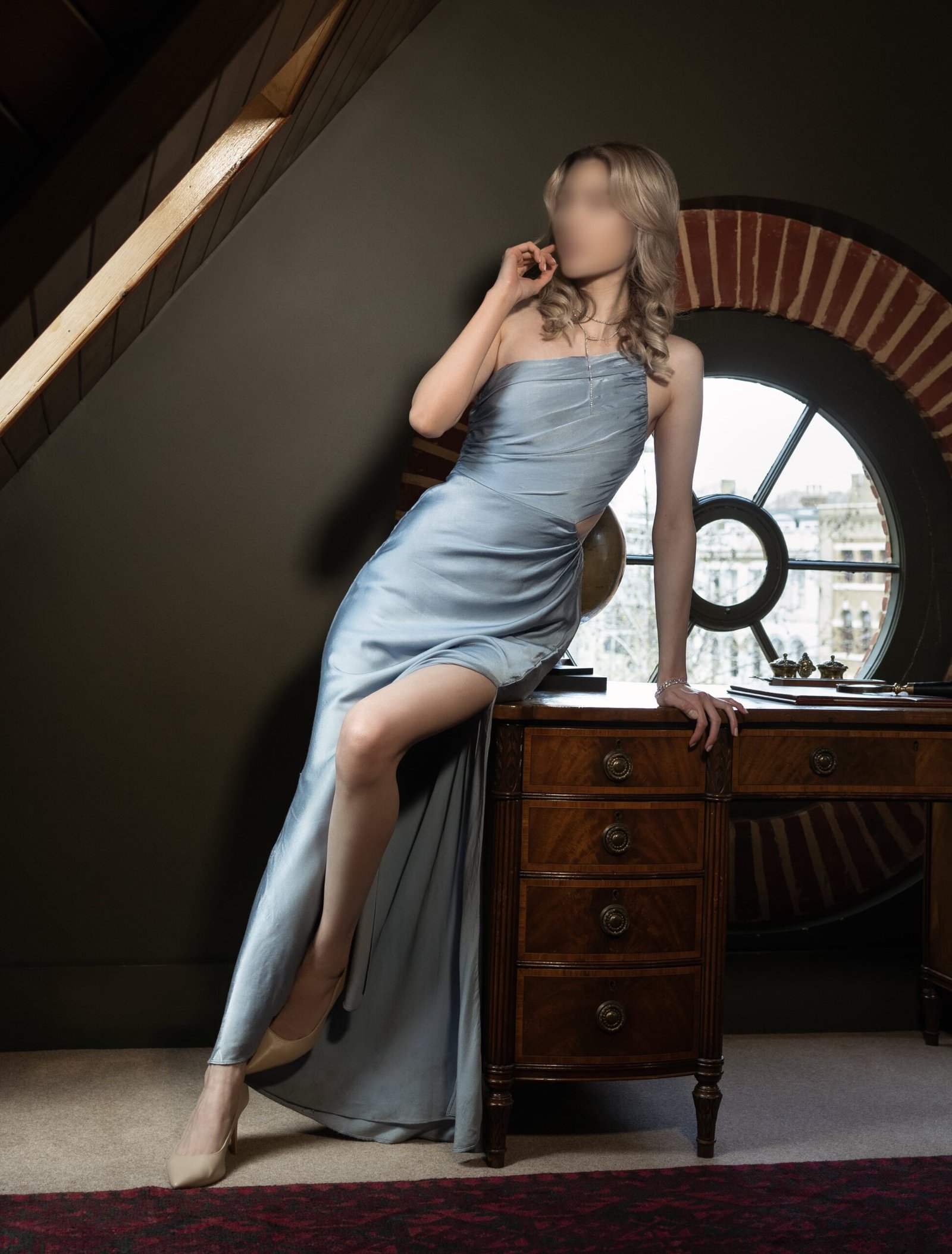 A blonde woman sits elegantly at an antique desk, their posture relaxed and contemplative. They are wearing a stylish, one-shoulder, sky-blue gown with a high leg slit, paired with beige heels. The room is characterized by its classic architecture, including a round port-style window that offers a glimpse of the urban landscape outside, melding historical charm with a touch of contemporary fashion.