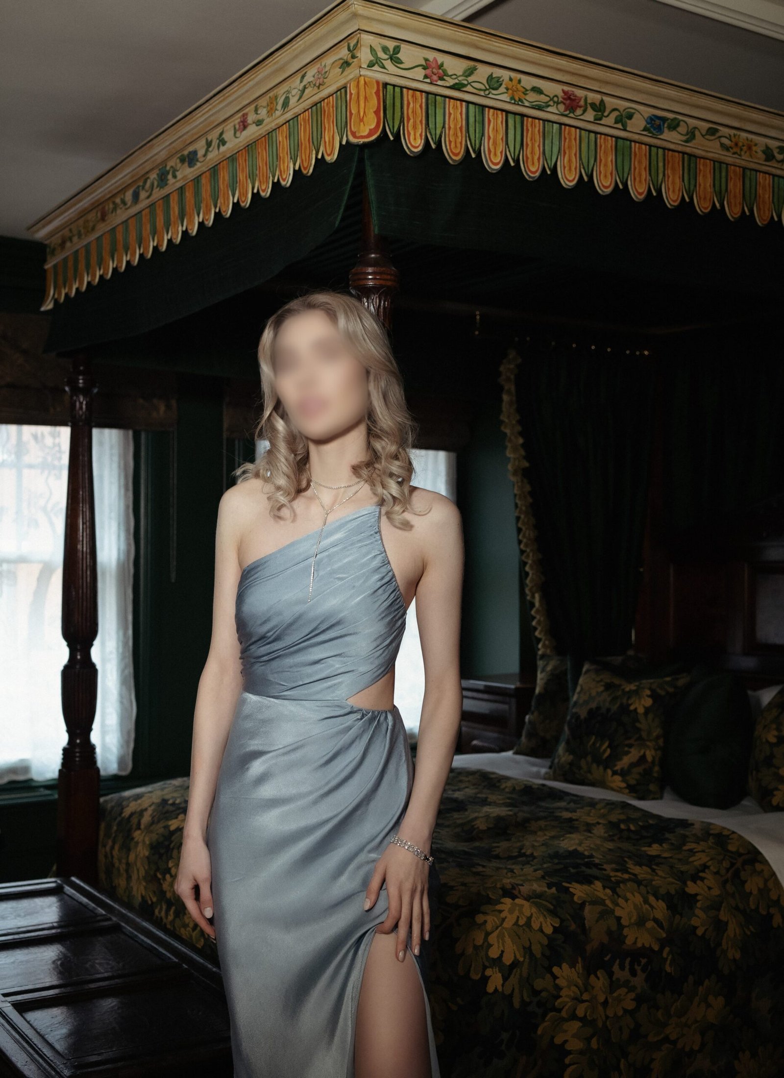 A young blonde woman standing in an opulent room with a classic four-poster bed adorned with a decorative green and floral canopy. They are dressed in an elegant, one-shoulder, silvery-blue gown with a tasteful side cut-out, a thigh-high slit, and are accessorized with a delicate necklace and bracelet. The room exudes an air of historical luxury, with dark wood furnishings and deep green drapery enhancing the stately ambiance.